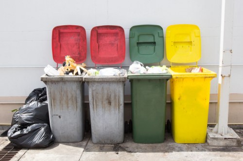 A row of four wheelie bins in different colours—grey, grey, green, and yellow—are positioned outdoors on a paved surface against a plain white wall. The lids of the grey bins are open, revealing assorted waste materials such as plastic bags, cardboard, and miscellaneous rubbish inside. To the left of the grey bins, there are several tightly tied black rubbish bags stacked on the ground. The green and yellow bins are closed, with their lids also slightly ajar, and appear to be used for different waste types, consistent with a commercial or community waste collection area. The bins are made of durable plastic, showing signs of regular use, with some dirt or grime visible on their surfaces. The environment suggests a designated waste disposal point, typical of premises in Hemel Hempstead, where Commercial Waste Hemel Hempstead provides rubbish removal services. Natural daylight illuminates the scene, emphasizing the textures of the plastic bins and the waste materials, depicting a typical urban waste management setup on a residential or commercial frontage in the town.