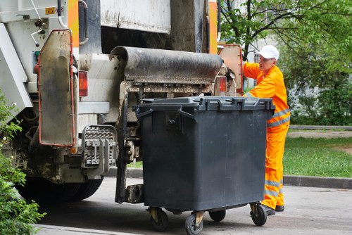 A waste collection operative dressed in bright orange high-visibility workwear, including trousers and a cap, is standing next to a black wheeled rubbish bin positioned in front of a large industrial waste collection vehicle on a paved street. The bin has a textured plastic surface, with visible handles and a lid that is closed. The vehicle features a metal hopper with a worn, textured surface, and mechanisms for lifting and emptying bins, painted in weathered grey and orange tones. The background includes greenery and trees, suggesting a suburban or residential area, with natural daylight illuminating the scene. The operative appears to be engaged in the process of waste collection, with a focus on rubbish removal services that might operate in Hemel Hempstead or nearby regions. This image underscores the professionalism and safety measures involved in commercial waste management, as carried out by companies like Commercial Waste Hemel Hempstead, supporting local rubbish disposal needs.