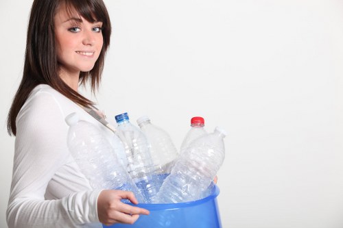A young woman with shoulder-length brown hair, wearing a white long-sleeved top, is smiling slightly while holding a blue plastic bin filled with several clear plastic bottles, some with blue caps and others with red caps. The bottles are different sizes and appear to be empty, with some slightly crushed or bent. The background is plain and white, emphasizing the bottles and the woman. The scene suggests activities related to waste collection or recycling, which aligns with rubbish removal services offered by companies such as Commercial Waste Hemel Hempstead. The image's outdoor or indoor context is not specified, but the clean background keeps the focus solely on the woman and the bottles, highlighting the process of handling plastic waste, relevant to waste management and recycling efforts in the Hemel Hempstead area.