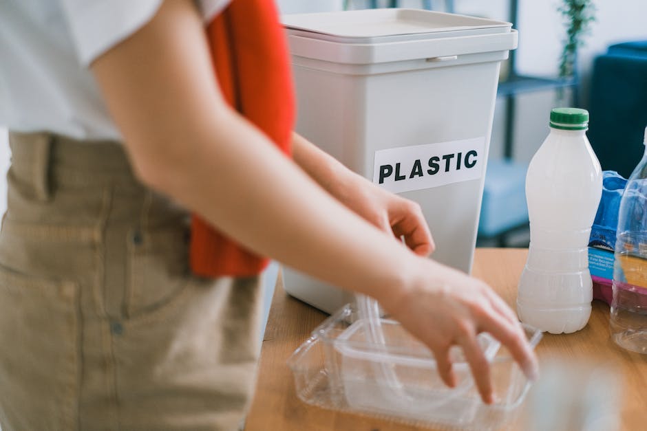 A person holding a clear plastic bag filled with empty transparent plastic bottles, with the bottle caps visible and some bottles having ridged necks. The bag appears to be made of thin, flexible plas