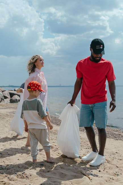 A man with dark skin dressed in a red t-shirt, light blue denim shorts, white sneakers, and a black cap, is standing on a sandy beach holding a large white rubbish bag. Beside him, a young boy wearing