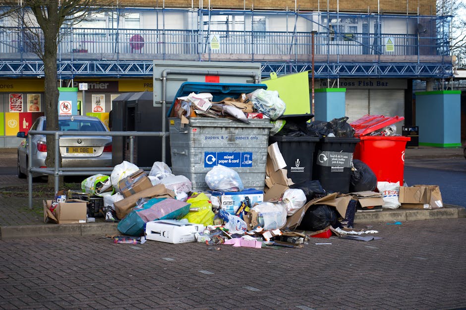 A tall, bright blue wheelie bin with a closed lid, positioned outdoors on a grassy area with sunlight illuminating the scene. The bin has a small, rectangular sticker on its front that reads 'Fight Ba